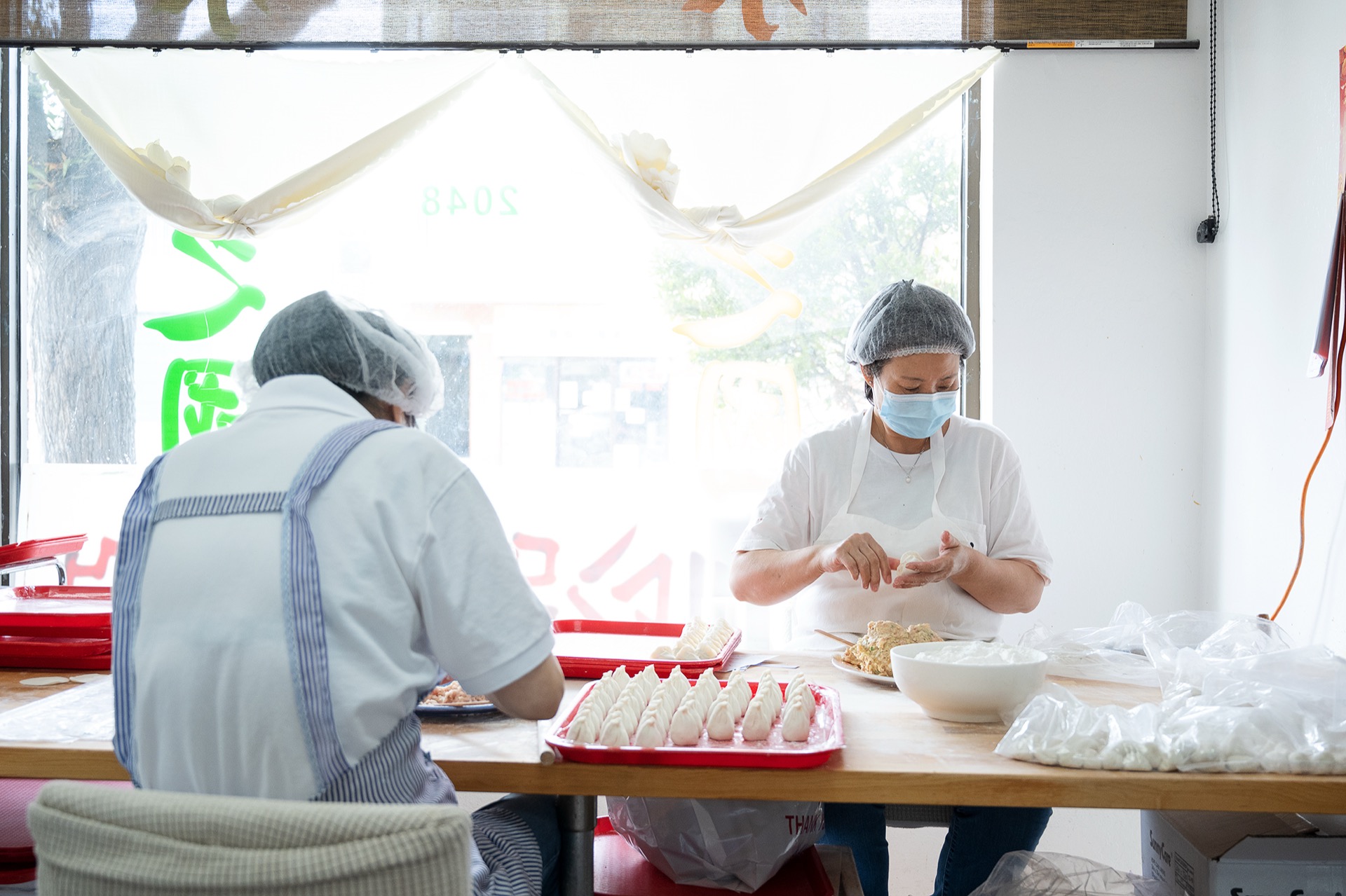 Two staff hand-folding dumplings at a sunlit table by the restaurant window
